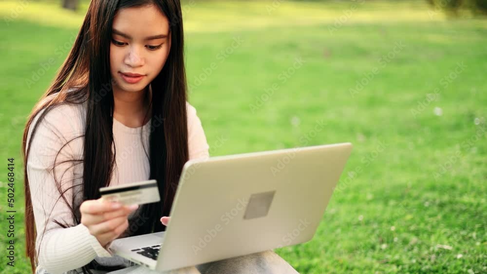 Young attractive asian woman paying with credit card on laptop sitting on the grass in the park. Happy customer. Young attractive asian woman paying with credit card on laptop sitting on the grass in the park. Happy customer.