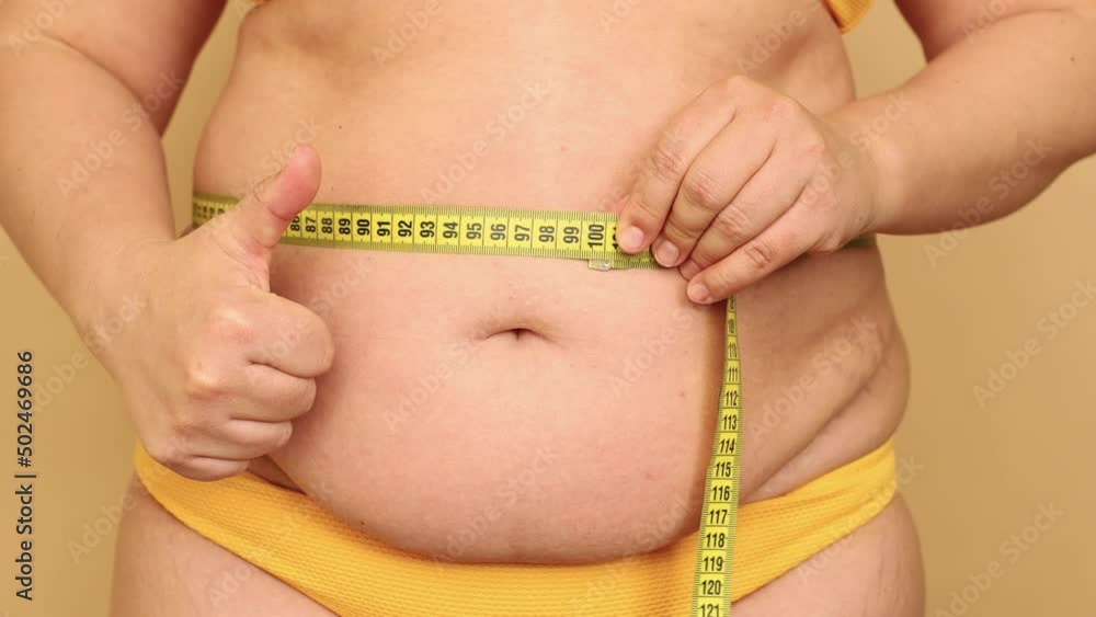 Close-up of unrecognizable fat woman standing in yellow bikini, showing thumb up sign, measuring waist with tape on beige background. Health problems, cellulite, obesity, weight control, liposuction. Close-up of unrecognizable fat woman standing in yellow bikini, showing thumb up sign, measuring waist with tape on beige background. Health problems, cellulite, obesity, weight control, liposuction.