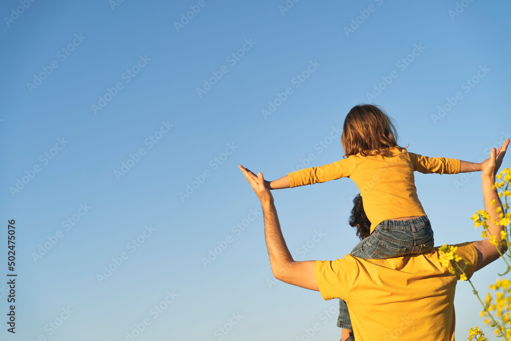 Dad holds his daughter on his shoulders. Portrait of a young father carrying his daughter on his back. father's day. Dad and son playing together outdoors on a summer. Family happiness love concept Dad holds his daughter on his shoulders. Portrait of a young father carrying his daughter on his back. father's day. Dad and son playing together outdoors on a summer. Family happiness love concept