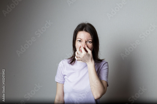 Portrait of a frustrated woman pinch her nose with disgust on her face because of a bad smell on a gray background
