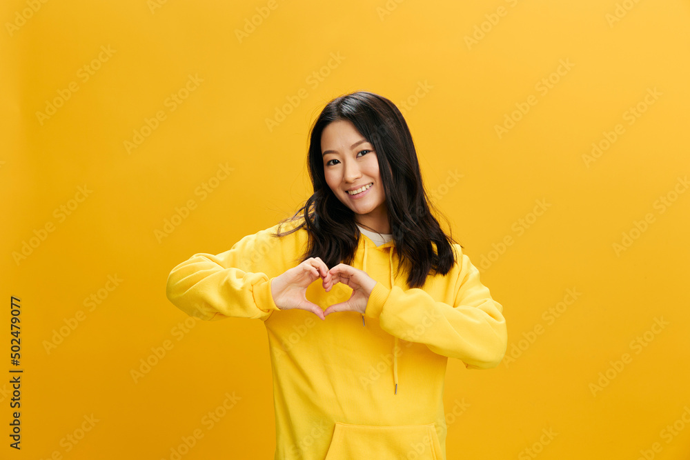 Lovely falling in love happy Asian student young woman in yellow hoodie sweatshirt show heart shaped sign posing isolated on over orange studio background. Cool offer. People Emotions concept Lovely falling in love happy Asian student young woman in yellow hoodie sweatshirt show heart shaped sign posing isolated on over orange studio background. Cool offer. People Emotions concept