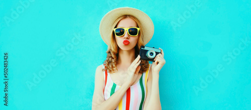 Summer portrait of happy stylish young woman photographer with film camera blowing her lips with red lipstick wearing straw hat on blue background, blank copy space for advertising text