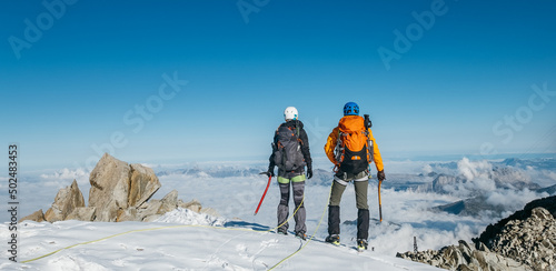 Couple connected team rope with climbing harness dressed mountaineering clothes with backpacks and ice axes enjoying views ascending Mont Blanc (Monte Bianco) summit near Aiguille du Midi, France.