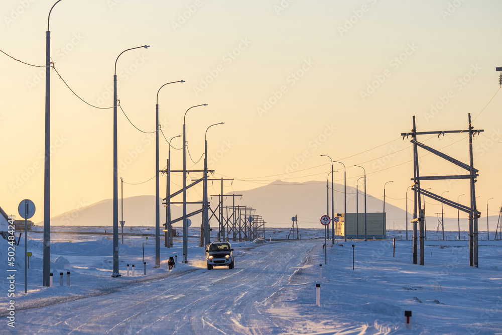Snowy road in the Arctic tundra. Winter sunset view of the road covered with snow and ice. A car is driving along the road. Transport infrastructure in the Far North. Chukotka, Polar Siberia, Russia.