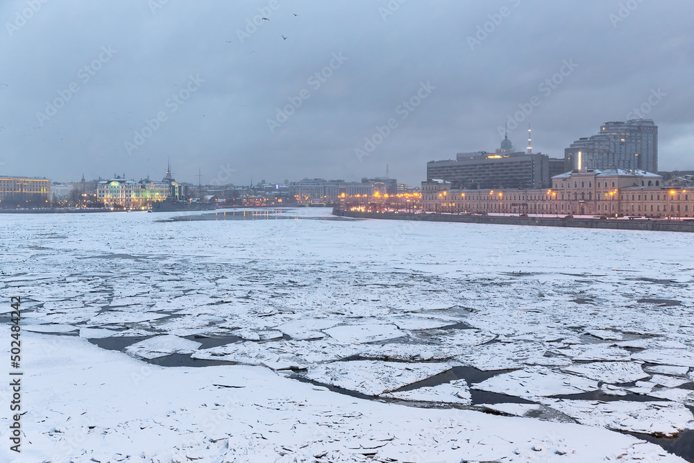 Winter view of the Neva River in the city center of St. Petersburg, Russia. Cracked ice floes on the surface of the water. Cold winter weather. Historical and modern buildings on the embankment.