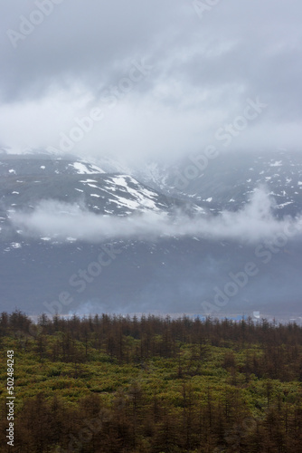 View of the mountains in the clouds. There is snow in the distance on the slopes of the mountains. Poor visibility in cloudy weather. Natural background. Magadan Region, Siberia, Far East Of Russia.