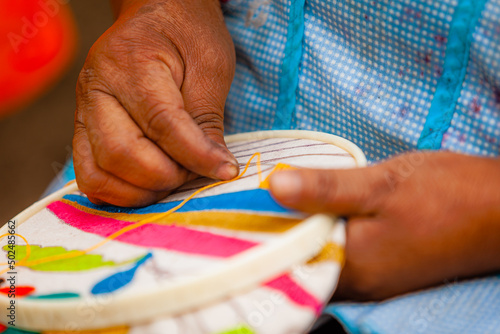 Extreme closeup photography of the hands of an indigenous woman embroidering a Tenango, traditional Mexican embroidery with orange thread and needle.