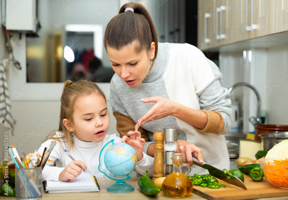 Positive mother cooking and little daughter doing school homework at kitchen