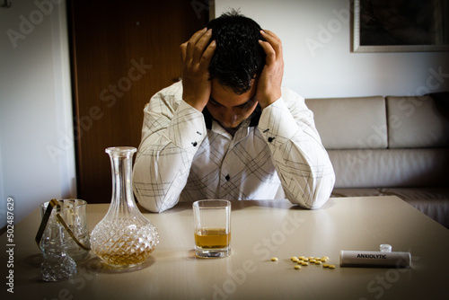 Desperate man with his hands on his head and leaning on the table at home after consuming psychiatric drugs and alcohol. photo
