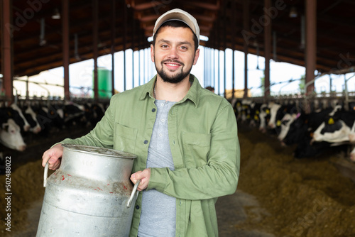 Vászonkép Closeup portrait of cheerful young bearded dairy farm owner carrying aluminum ca