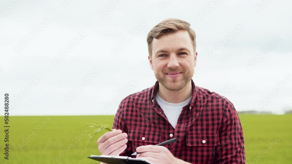 Portrait shot of attractive farmer standing in green field with green wheat and notebook in the hands. Farmer with smile outdoors in summer Portrait shot of attractive farmer standing in green field with green wheat and notebook in the hands. Farmer with smile outdoors in summer