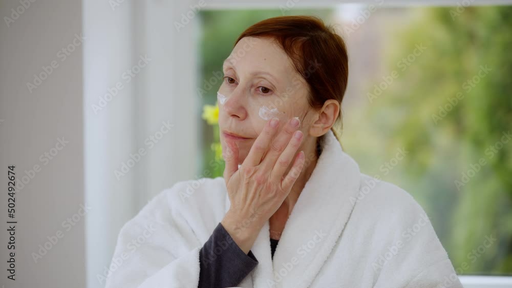 Redhead mature woman applying facial moisturizer looking away. Portrait of Caucasian retiree in white bathrobe at home in the morning. Domestic life and beauty routine concept