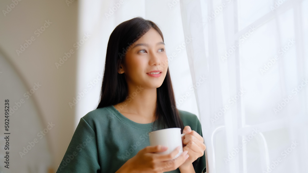 Young asian woman standing beside window and holding mug in bedroom at home, She drinking milk after wake up in the morning Young asian woman standing beside window and holding mug in bedroom at home, She drinking milk after wake up in the morning