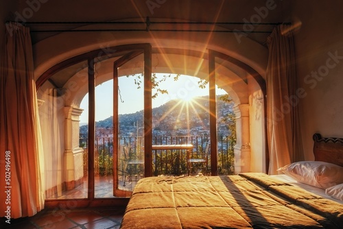 Outside the windows of the hotel is a city on the slopes of the mountains. The rays of the evening sun and the roofs of houses. Balcony with chairs, glass doors, beds and room interior with colonial a