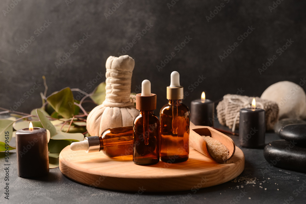 Wooden board with bottles of essential oil, sea salt, massage bag and burning candles on dark background - obrazy, fototapety, plakaty 