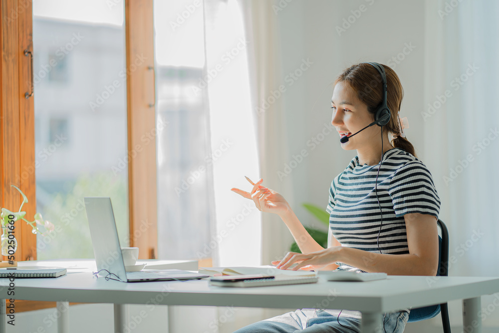 Young Asian student wearing headphones using a laptop Write a note about learning a language. View webinars online. audio listening course e-learning concept Young Asian student wearing headphones using a laptop Write a note about learning a language. View webinars online. audio listening course e-learning concept