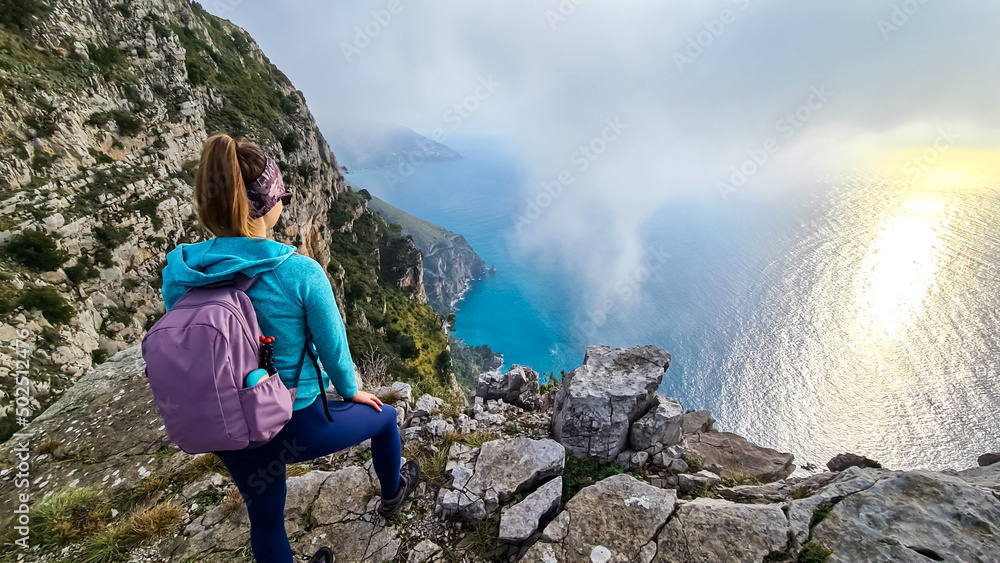 Naklejka premium Woman with scenic view from a hiking trail on the coastal driving road of beautiful Amalfi Coast, Campania, Italy, Europe. Riviera coastline at Mediterranean sea. Panoramic curvy road near Positano