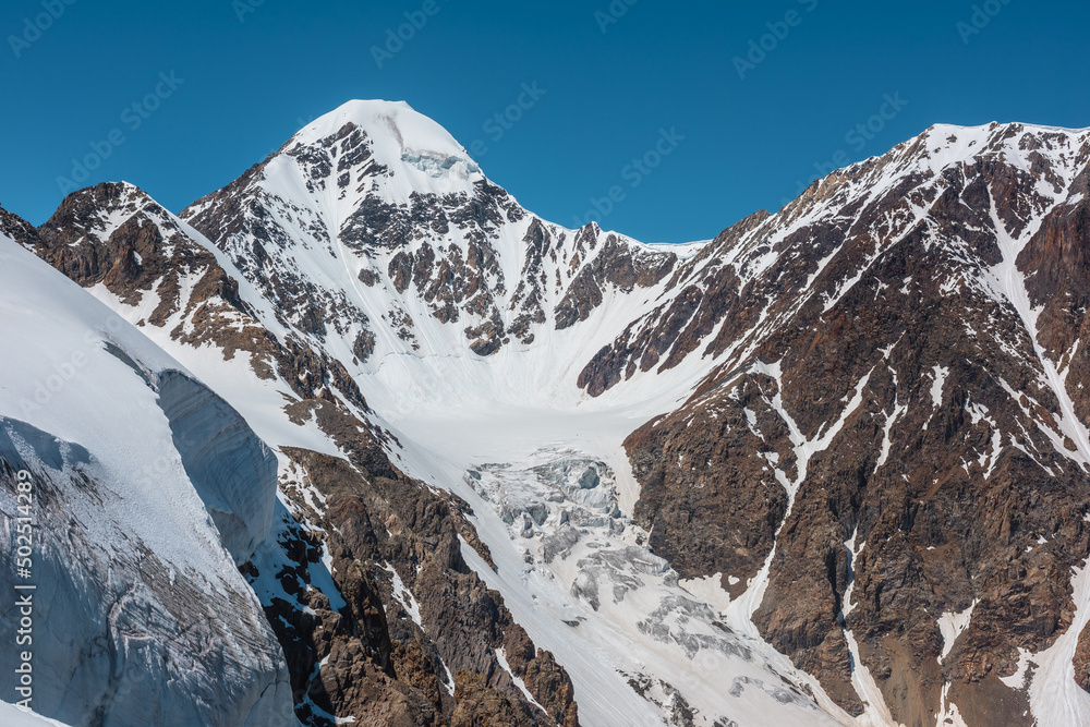 Scenic alpine landscape with snow-covered mountain top and steep glacier tongue with icefall in sunlight. Awesome mountain scenery with pointed peak and glacier on rocks under blue sky in sunny day. Scenic alpine landscape with snow-covered mountain top and steep glacier tongue with icefall in sunlight. Awesome mountain scenery with pointed peak and glacier on rocks under blue sky in sunny day.