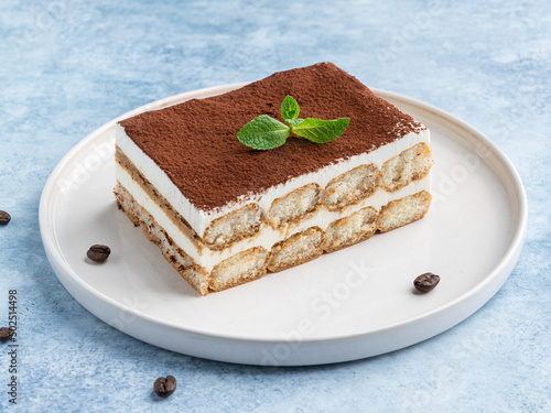 Tiramisu cake decorated with cocoa powder and fresh green mint leaf. Close up food. Blue background. Traditional italian dessert. Piece of cake on white ceramic plate.