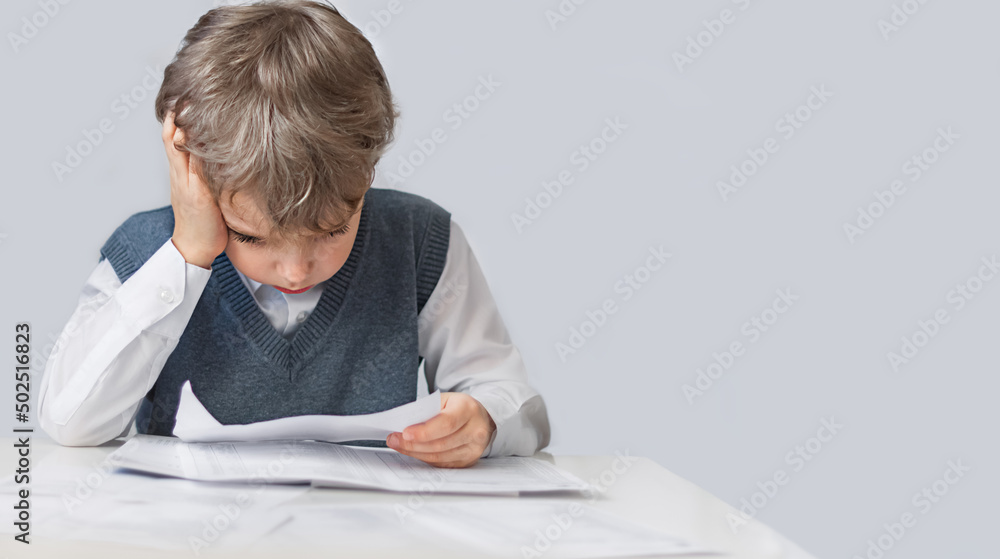 a little boy sits at a table, holds a sheet in his hand and looks at it thoughtfully. Learning concept a little boy sits at a table, holds a sheet in his hand and looks at it thoughtfully. Learning concept