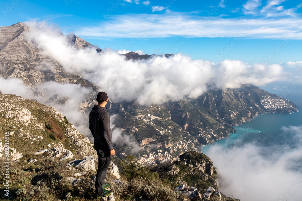 Happy man standing at cliff with scenic view from Monte Comune on the coastal town Positano. Magical hiking above clouds in Lattari Mountains, Apennines, Amalfi Coast, Campania, Italy, Europe. Awe