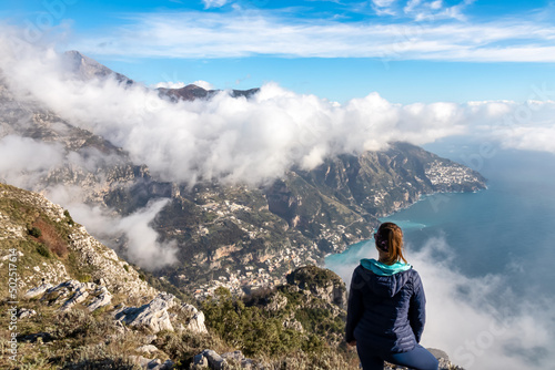 Happy woman standing at cliff with scenic view from Monte Comune on the coastal town Positano. Magical hiking above clouds in Lattari Mountains, Apennines, Amalfi Coast, Campania, Italy, Europe. Awe