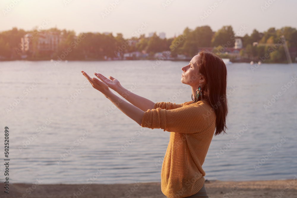 Young woman raises her hands to setting sun on shore lake, in yellow sweater enjoys life. Happy caucasian girl is enjoying outdoors near the water.