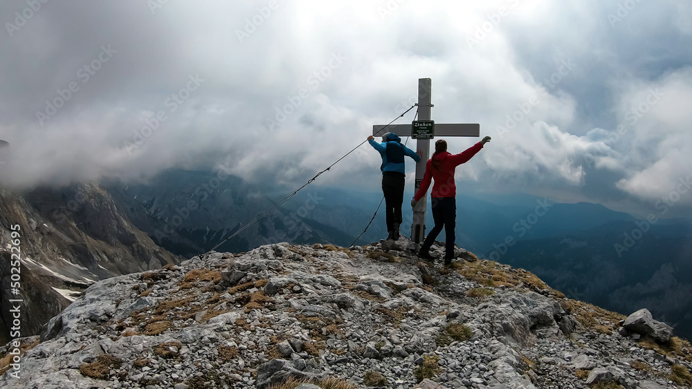 Active couple on the cloud covered summit cross of mount Zinken in the Hochschwab Region in Upper Styria, Austria. On top of a mountain peak in the Alps in Europe. Freedom fun vibe. Rainy dark clouds