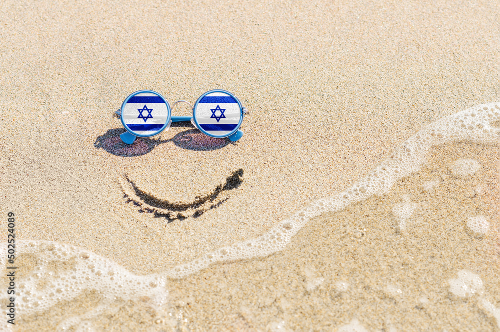 A painted smile on the beach and sunglasses with the flag of Israel. The concept of a positive holiday in the resort of Israel.