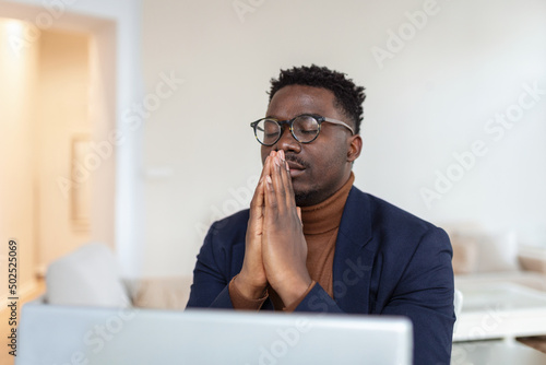 African guy sit at desk with clasped palms closed eyes thinking search problem solution, collect thoughts together, organize his emotional state before important meeting. Prayer hands sign of ask help