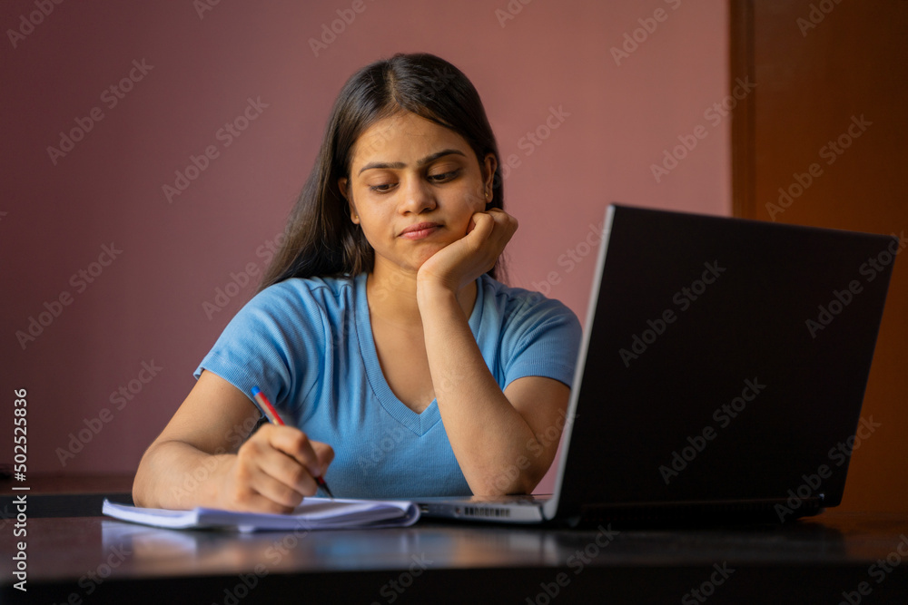 Indian girl sitting at the desk studying or working on laptop making notes Indian girl sitting at the desk studying or working on laptop making notes