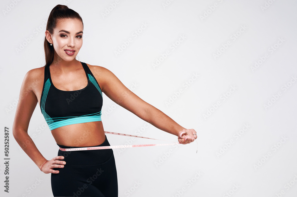 Strong fitness female in sport suit measuring waist on gray isolated background Strong fitness female in sport suit measuring waist on gray isolated background