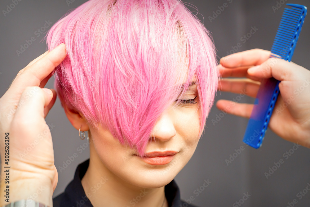 Fototapeta premium Hairdresser with hands and comb is checking out and fixing the short pink hairstyle of the young white woman in a hair salon