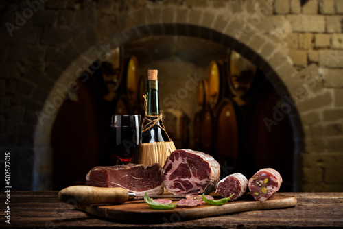 Flask of red wine with fresh smoked meat on rustic table with a cellar on background
