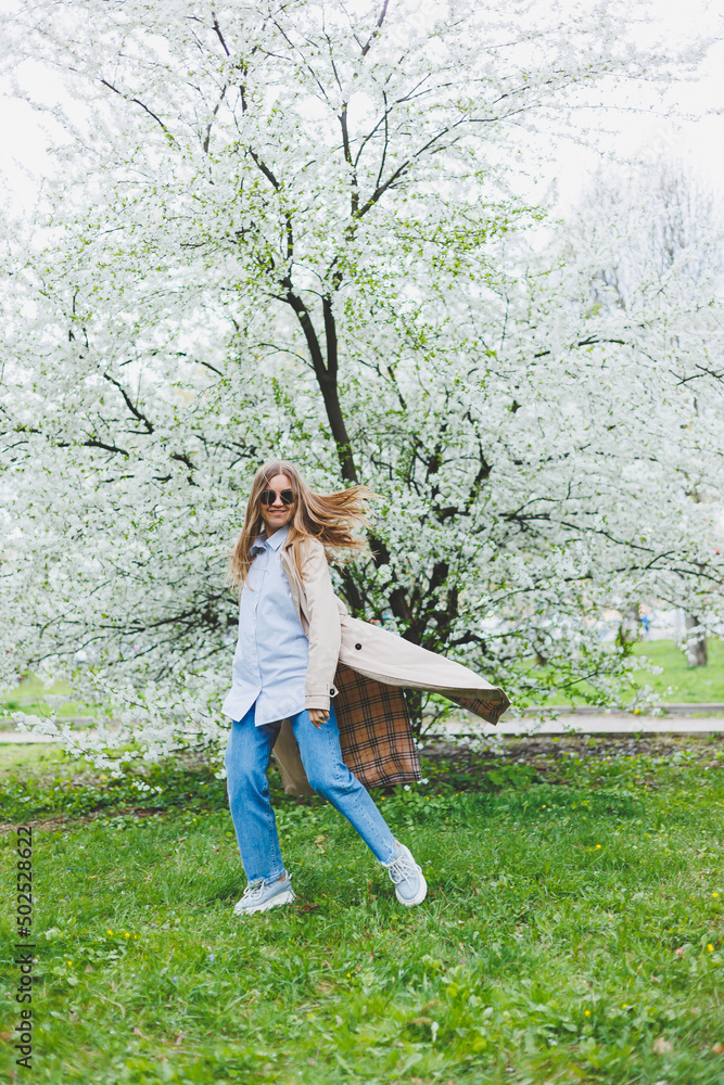 Naklejka premium A young woman with blond hair enjoys a blooming spring garden. Travel, spring break. Fashionable style. A woman in sunglasses and a beige trench coat runs through a flowering park. Selection focus