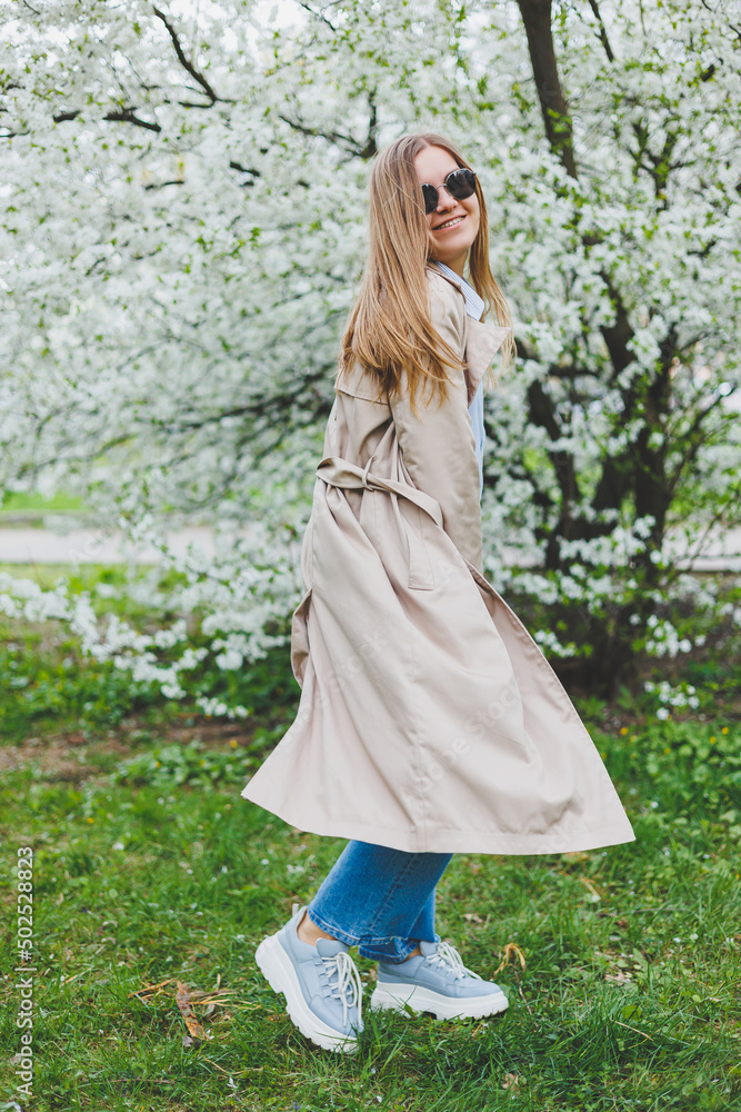 A young woman with blond hair enjoys a blooming spring garden. Travel, spring break. Fashionable style. A woman in sunglasses and a beige trench coat runs through a flowering park. Selection focus A young woman with blond hair enjoys a blooming spring garden. Travel, spring break. Fashionable style. A woman in sunglasses and a beige trench coat runs through a flowering park. Selection focus