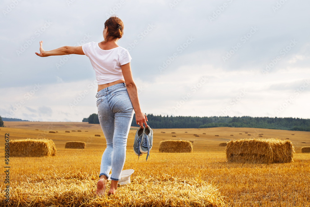 barefoot girl with sneakers in hand stands on a haystack on a bale in the agricultural field after harvesting