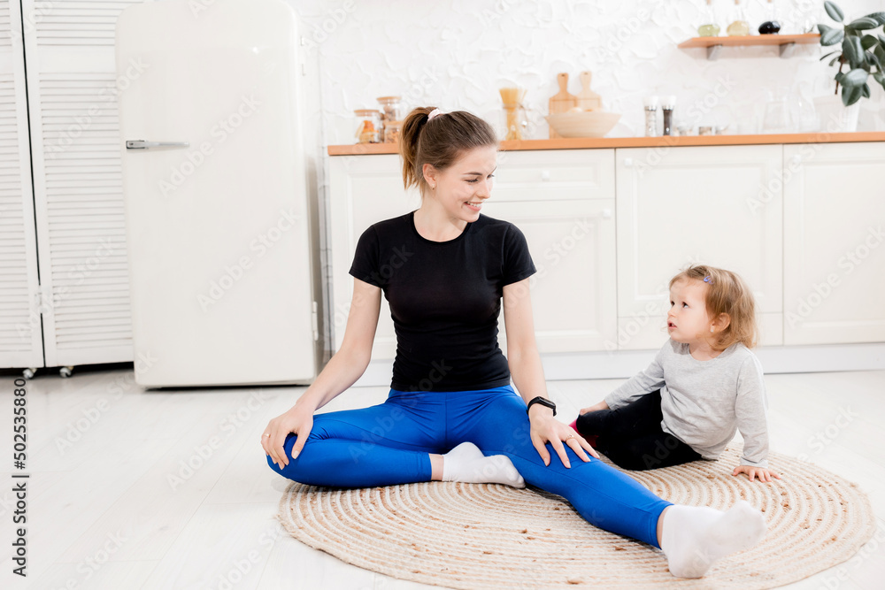Sporty brunette mother and daughter sitting on the kitchen, stretching and smiling, home interior. Happy female parent and girl kid in sportswear having moring training together in living room Sporty brunette mother and daughter sitting on the kitchen, stretching and smiling, home interior. Happy female parent and girl kid in sportswear having moring training together in living room