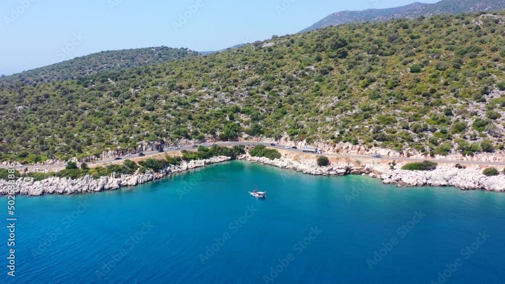 Wide aerial of a sailboat anchored in the tropical blue Mediterranean Sea near Finike Turkey alongside a green hill and coastal highway road Wide aerial of a sailboat anchored in the tropical blue Mediterranean Sea near Finike Turkey alongside a green hill and coastal highway road