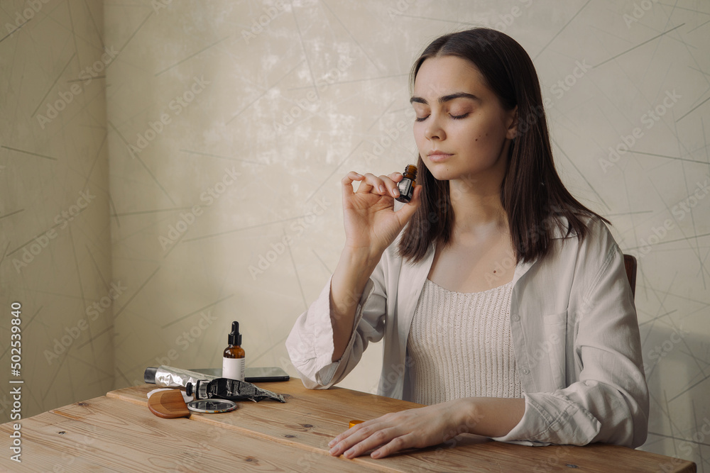 Relaxed woman with closed eyes smelling aromatic fragrance of natural essential oil from glass bottle while enjoying aromatherapy at table at home  - obrazy, fototapety, plakaty 