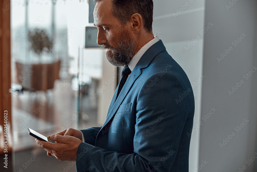Side view of businessman in stylish suit standing and using smartphone indoors Side view of businessman in stylish suit standing and using smartphone indoors