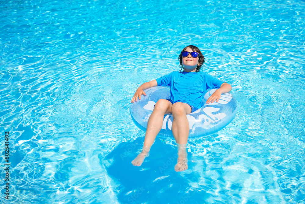 Boy wearing sun glasses, relaxing on an inflatable swim ring in a sunny day, spending time in a swimming pool, aquapark resort hotel. Summer holidays concept.