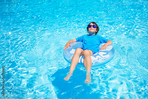Boy wearing sun glasses, relaxing on an inflatable swim ring in a sunny day, spending time in a swimming pool, aquapark resort hotel. Summer holidays concept.