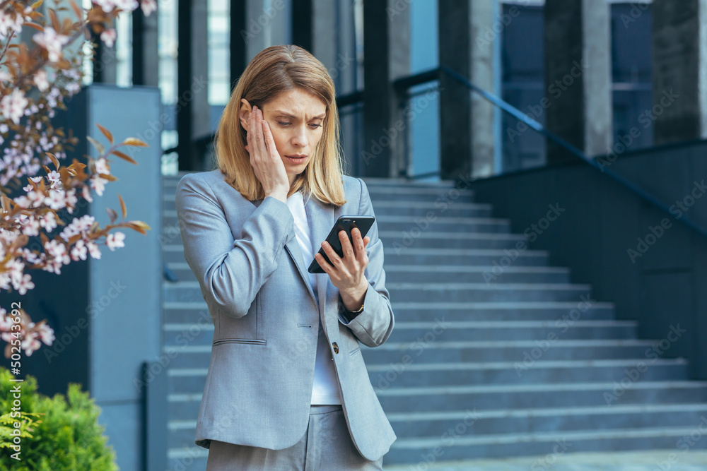 Frustrated and shocked business woman reading bad news online from phone, businesswoman outside office in business suit Frustrated and shocked business woman reading bad news online from phone, businesswoman outside office in business suit
