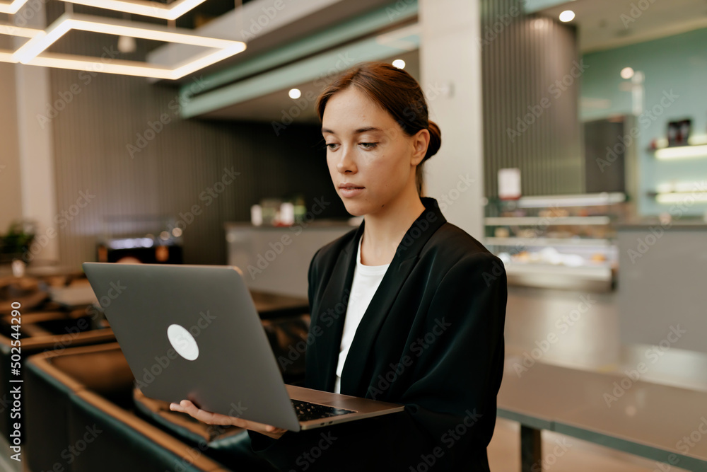 Tender caucasian woman in dark jacket with smile working in laptop and posing on background of modern office Tender caucasian woman in dark jacket with smile working in laptop and posing on background of modern office