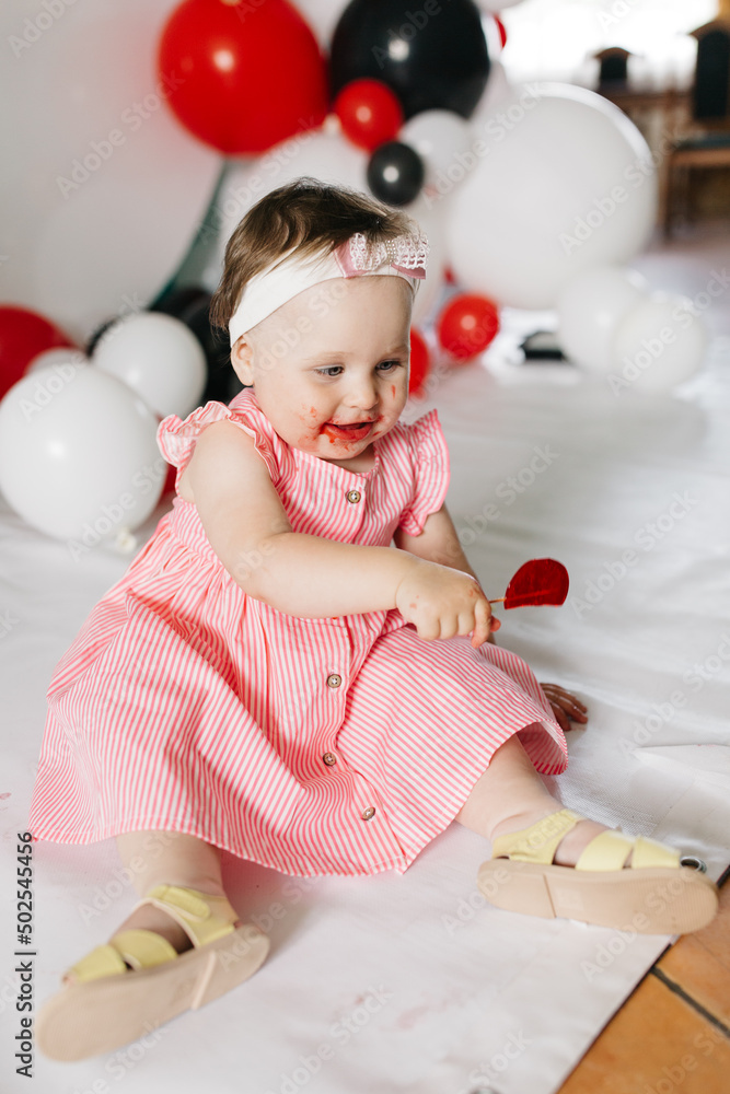 beautiful little one-year-old girl in a pink dress eats a lollipop. the child smeared himself with candy. kids love sweets