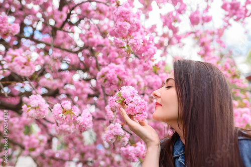 Young beautiful girl in a blue denim jacket sniffs the color of pink blossoming sakura. High quality photo
