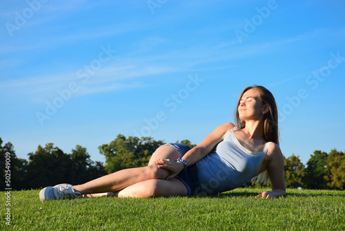 Beautiful girl with long hair in casual clothes laying on green lawn in summer park with blue sky enjoying sun rays. Summer, relax, calmness, leisure. Time to dream and fill with positive energy.