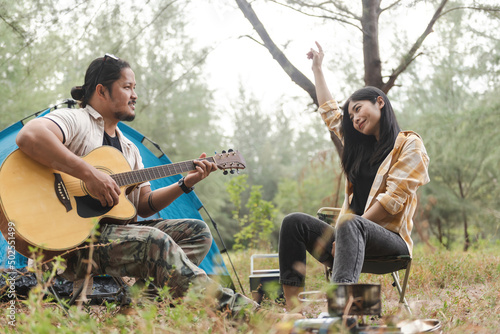 Asian couple playing guitar and singing while pitching a tent in the forest.