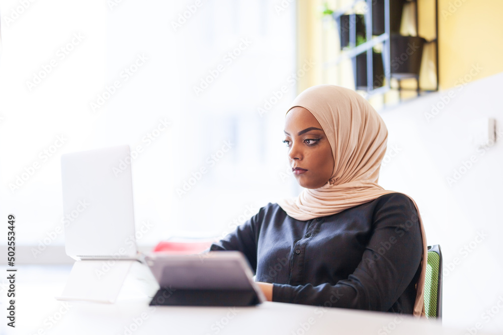 African american muslim girl with hijab working on a laptop in an office.. African american muslim girl with hijab working on a laptop in an office..